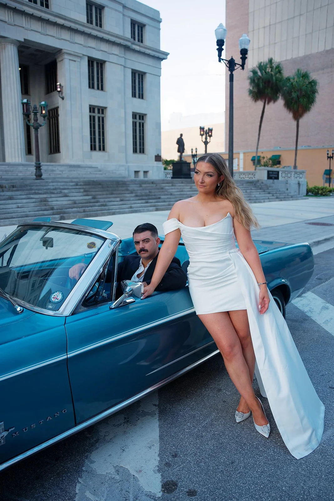 Rachel in a short white dress with a long train standing beside a vintage blue Mustang convertible with Austin in the driver's seat, downtown Miami courthouse behind them