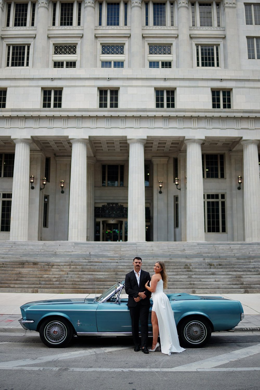 Rachel and Austin standing beside a blue vintage Mustang in front of the tall columns of the Miami-Dade County Courthouse