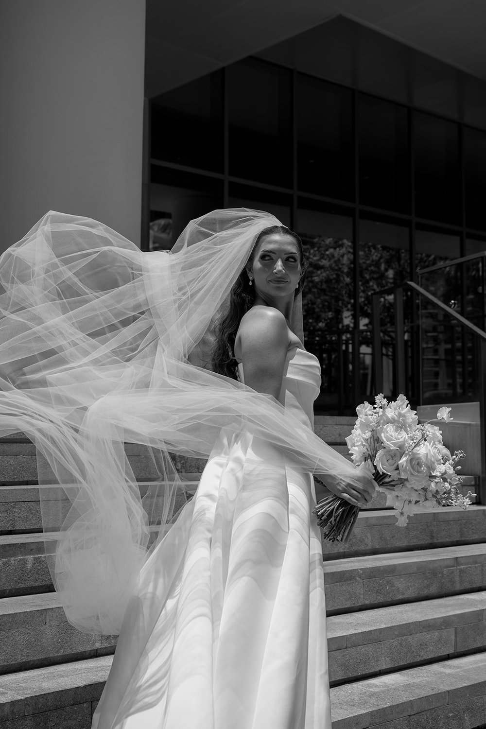 Rachel on stone steps with her veil blowing in the wind and a bouquet in her hand, in black and white