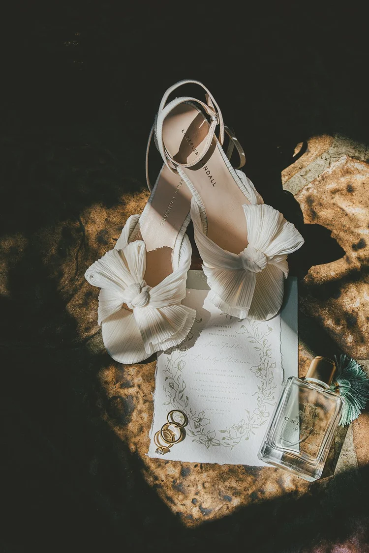 Film photo of white bow wedding shoes, wedding rings and a printed card in a patch of sunlight