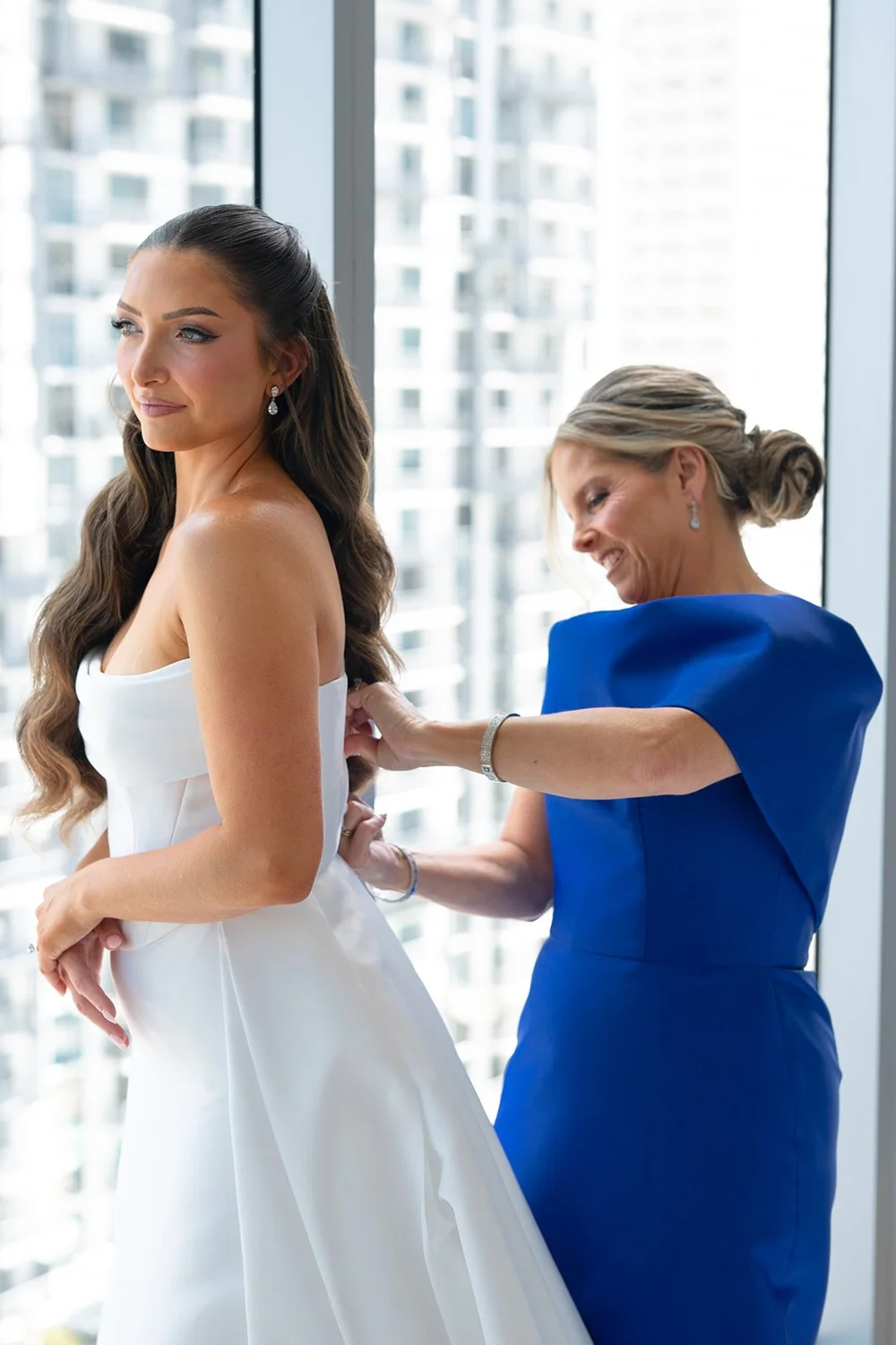 Rachel in her strapless gown at a window while a woman in a blue dress helps fasten it