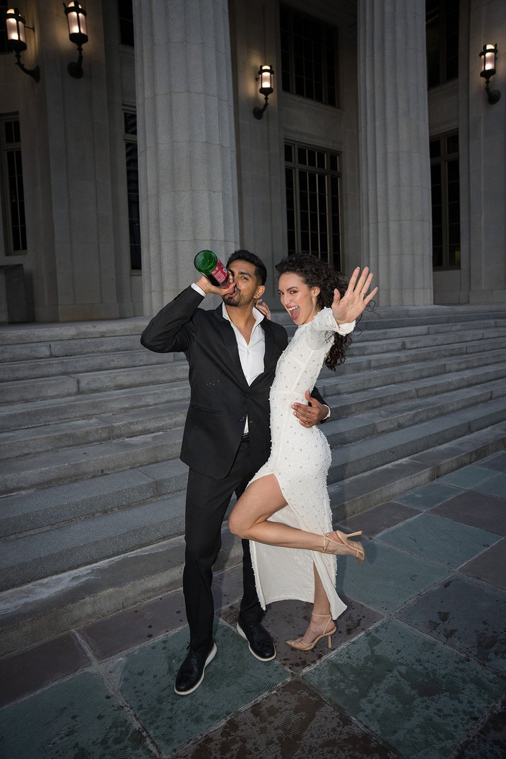 Bride kicking her leg up in a white dress on the courthouse steps at dusk while the groom lifts a bottle