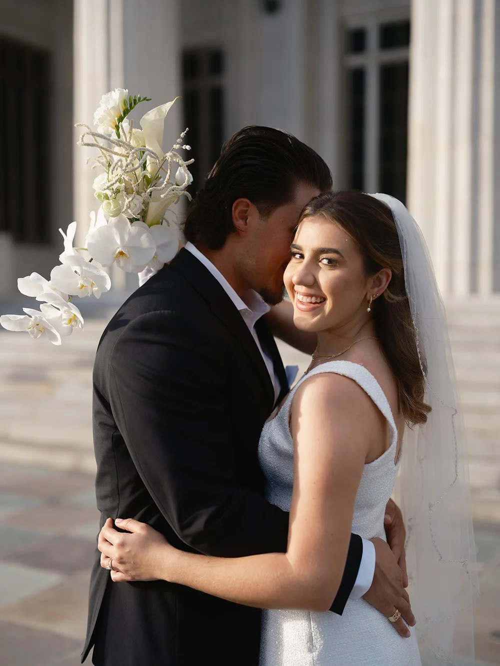 Groom holding the bride close, white flowers at the edge of the frame and her veil behind her