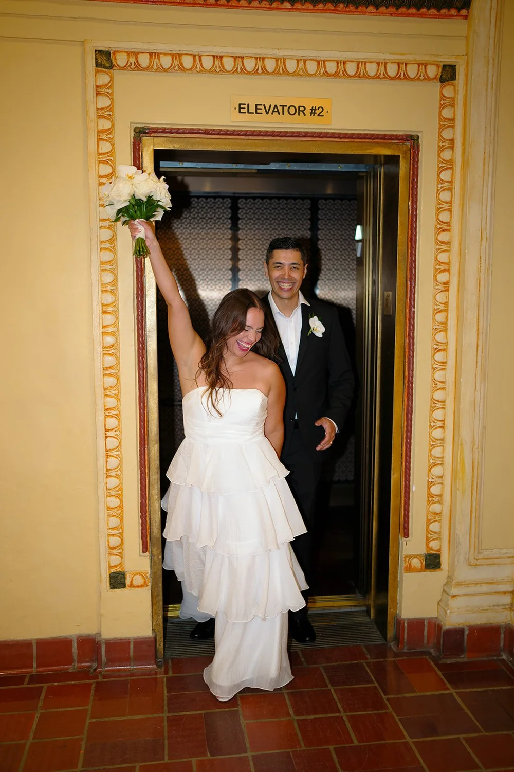 Chris lifting her white bouquet in the air in the elevator doorway with David behind her