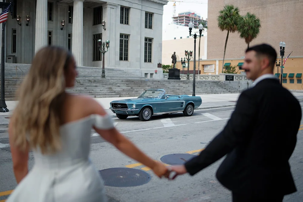 Rachel and Austin holding hands on a downtown Miami street with the blue Mustang in the distance