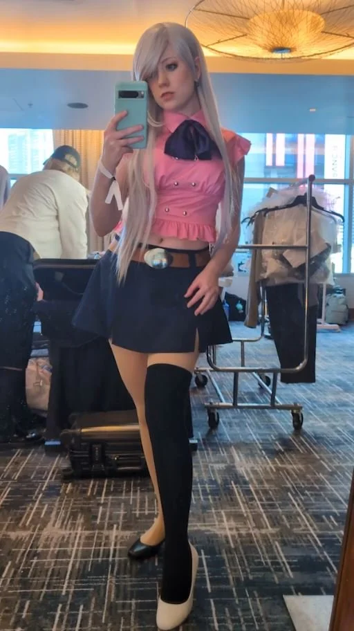Tiffany Gordon Cosplay as Elizabeth Liones from The Seven Deadly Sins, wearing a pink cropped top with a dark tie, a dark mini skirt with a green belt, and one long black stocking, posing in a forest.