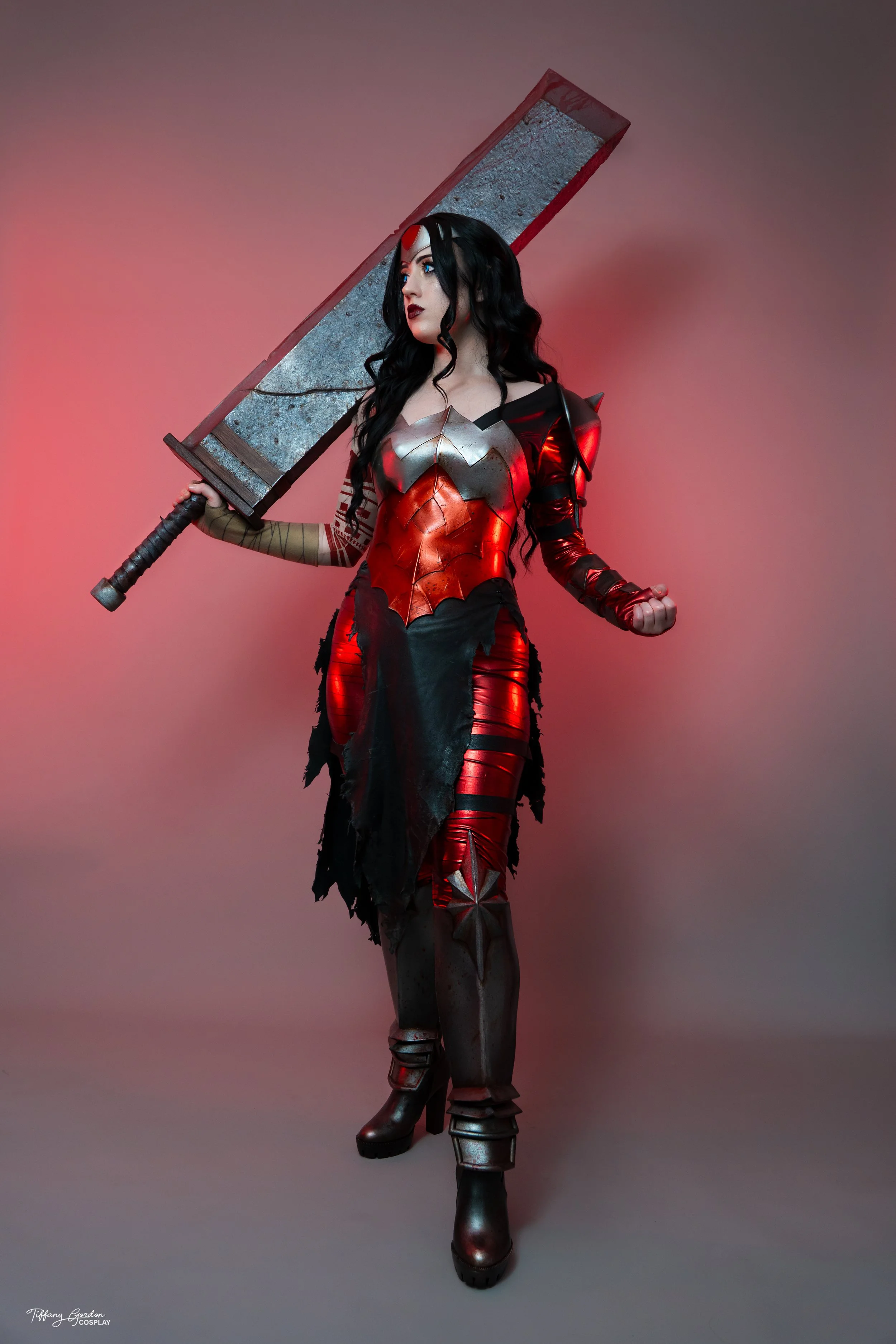 Full-body portrait of Tiffany Gordon’s Absolute Wonder Woman cosplay from DC Comics, featuring a red and black outfit with silver armor details, tattoo sleeve and long black hair holding her iconic sword.