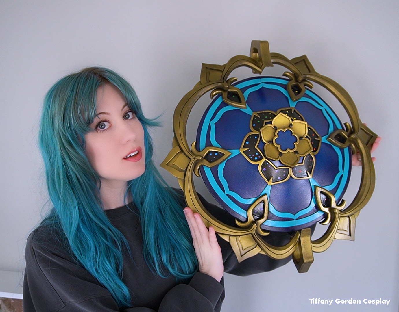 Tiffany Gordon Cosplay with blue hair holding a large, handcrafted Wanderer's hat from Genshin Impact, featuring a dark blue dome with teal lotus patterns and a complex gold filigree outer ring.
