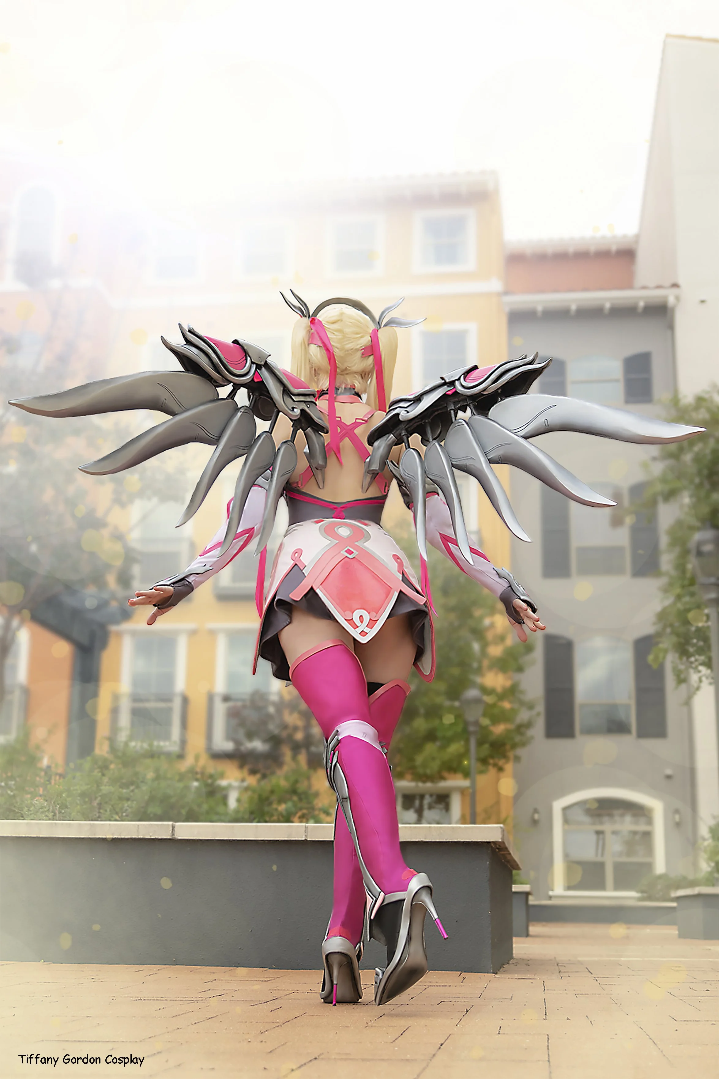 A back view shot of Tiffany Gordon Cosplay as Pink Mercy from Overwatch, showcasing the detailed silver and pink Valkyrie wings. The costume features the iconic pink ribbon symbol on the back of the skirt, vibrant pink thigh high boots.