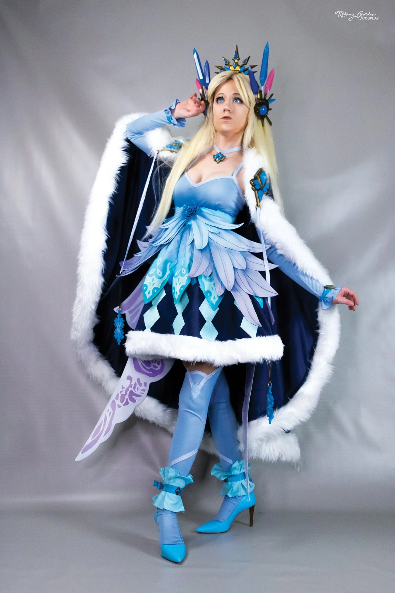 Andrea cosplay from Aura Kingdom by Tiffany Gordon Cosplay, featuring a blue feather-layered dress, a large white fur-lined cloak, and an intricate crystal crown.