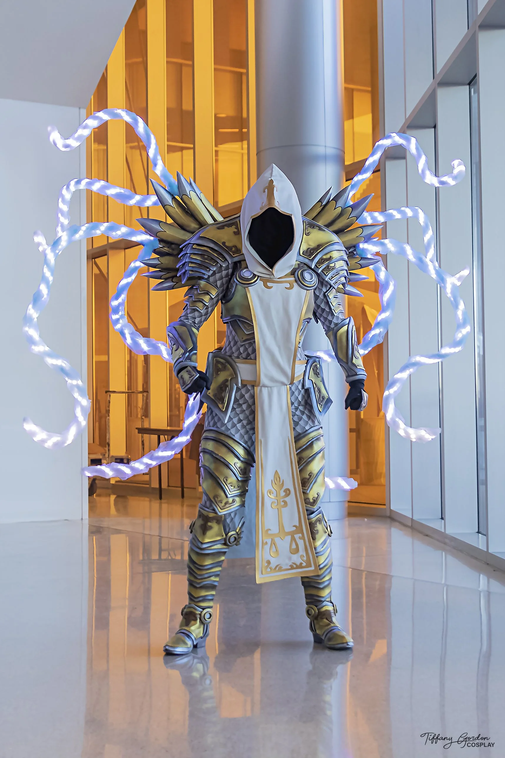 A full-body photo of Tiffany Gordon Cosplay's Tyrael cosplay standing in a modern hallway, featuring glowing blue LED wings, intricate gold and silver plate armor, and a white hooded tabard with gold embroidery.