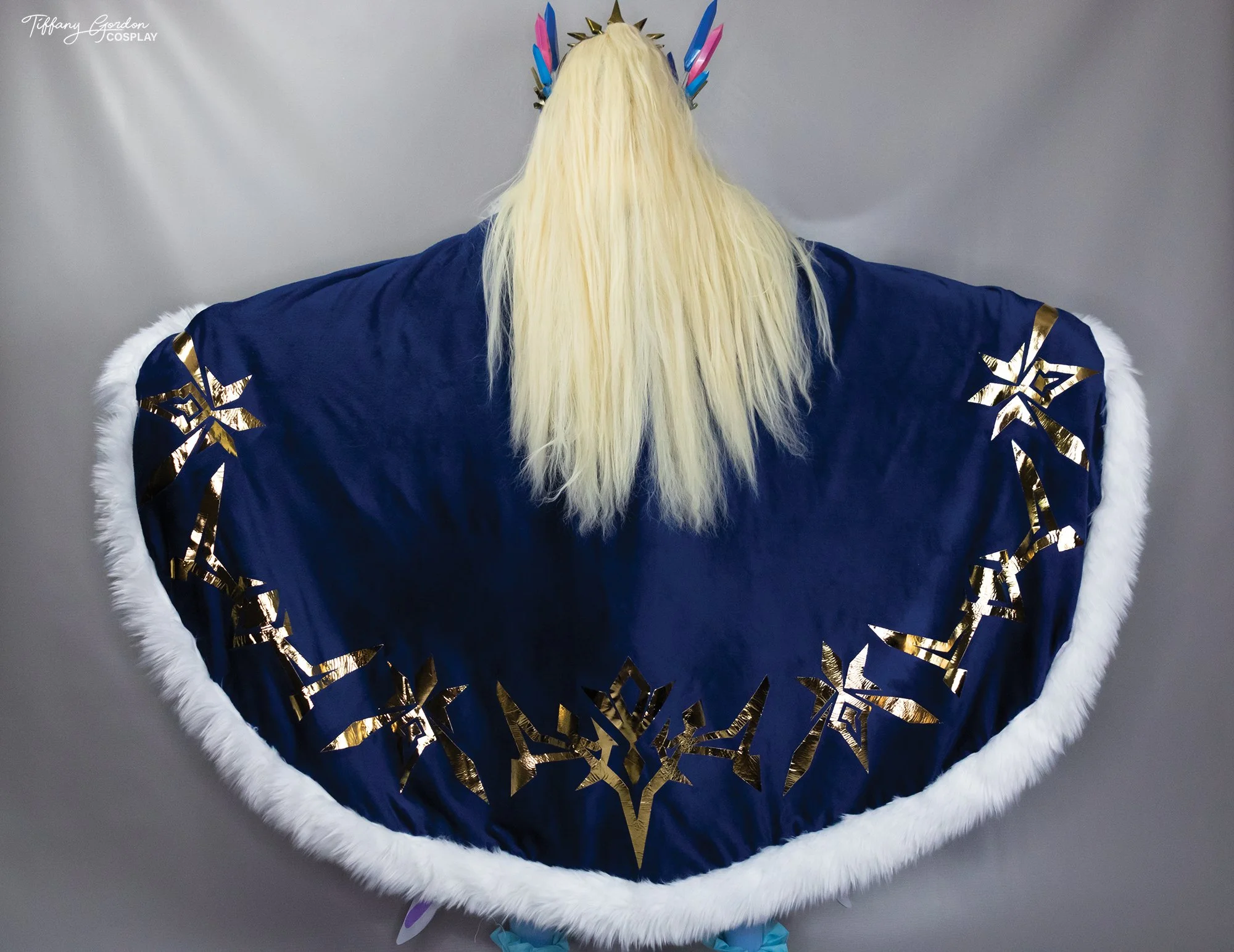 Rear view of Andrea cosplay from Aura Kingdom by Tiffany Gordon Cosplay, featuring a massive navy blue velvet cloak with gold star emblems, white fur trim, and long blonde hair.