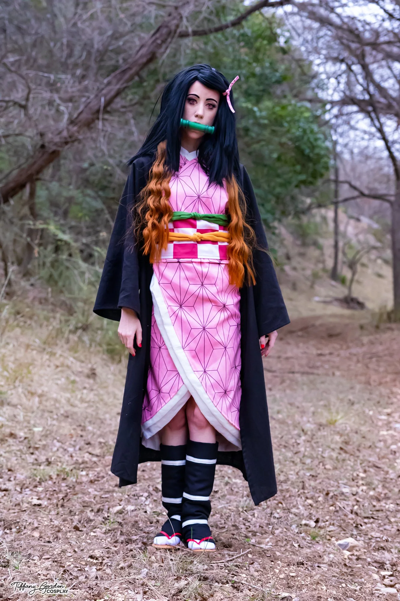 A full-body Nezuko Kamado cosplay from Demon Slayer by Tiffany Gordon Cosplay. The cosplayer is standing in a wooded path wearing a pink Asanoha-patterned kimono, a black haori, and the character's signature green bamboo muzzle and pink ribbon.