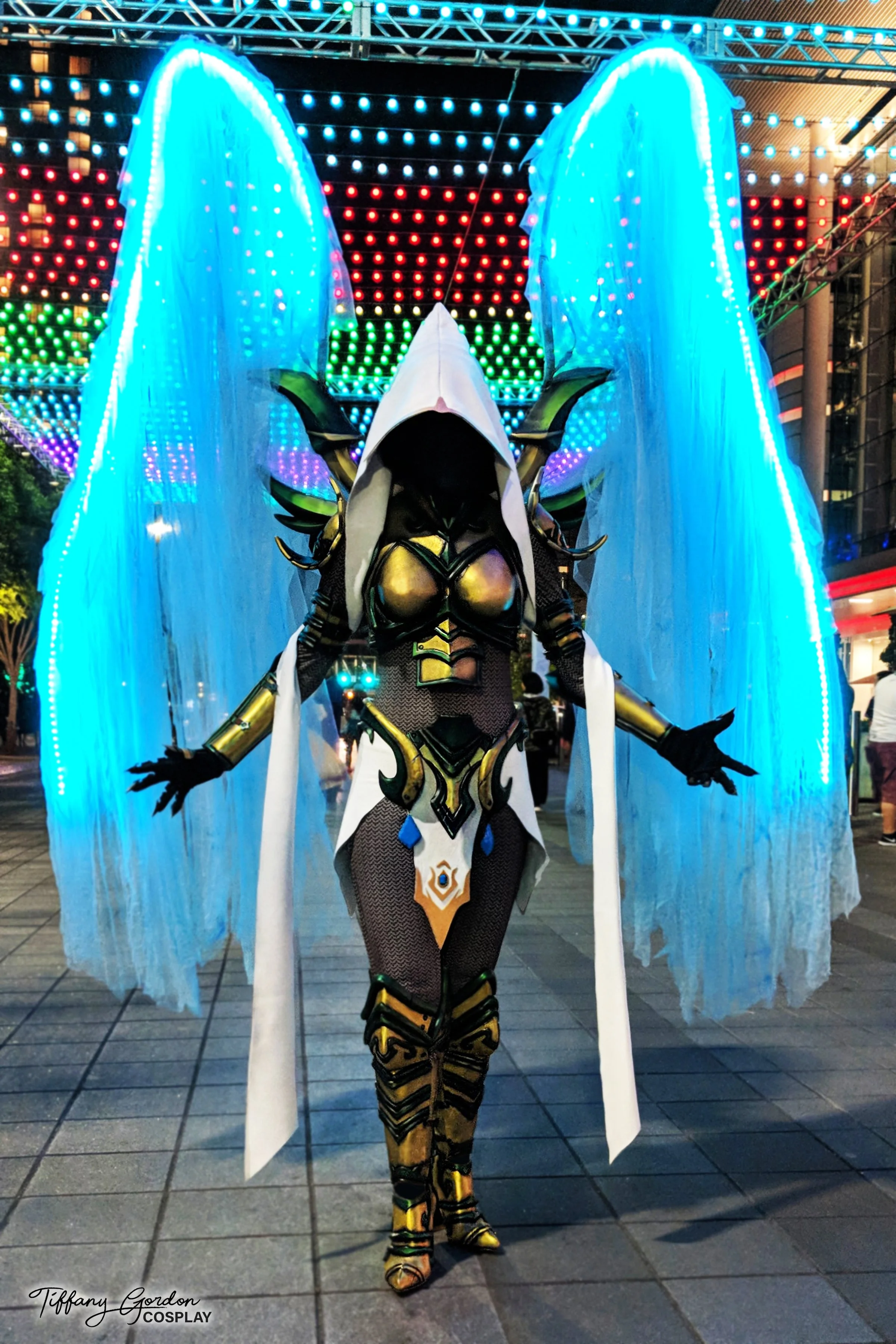 Auriel cosplay from Diablo by Tiffany Gordon Cosplay, featuring massive glowing blue LED wings, a white hooded cowl with a blacked-out face, and intricate gold and green plate armor.