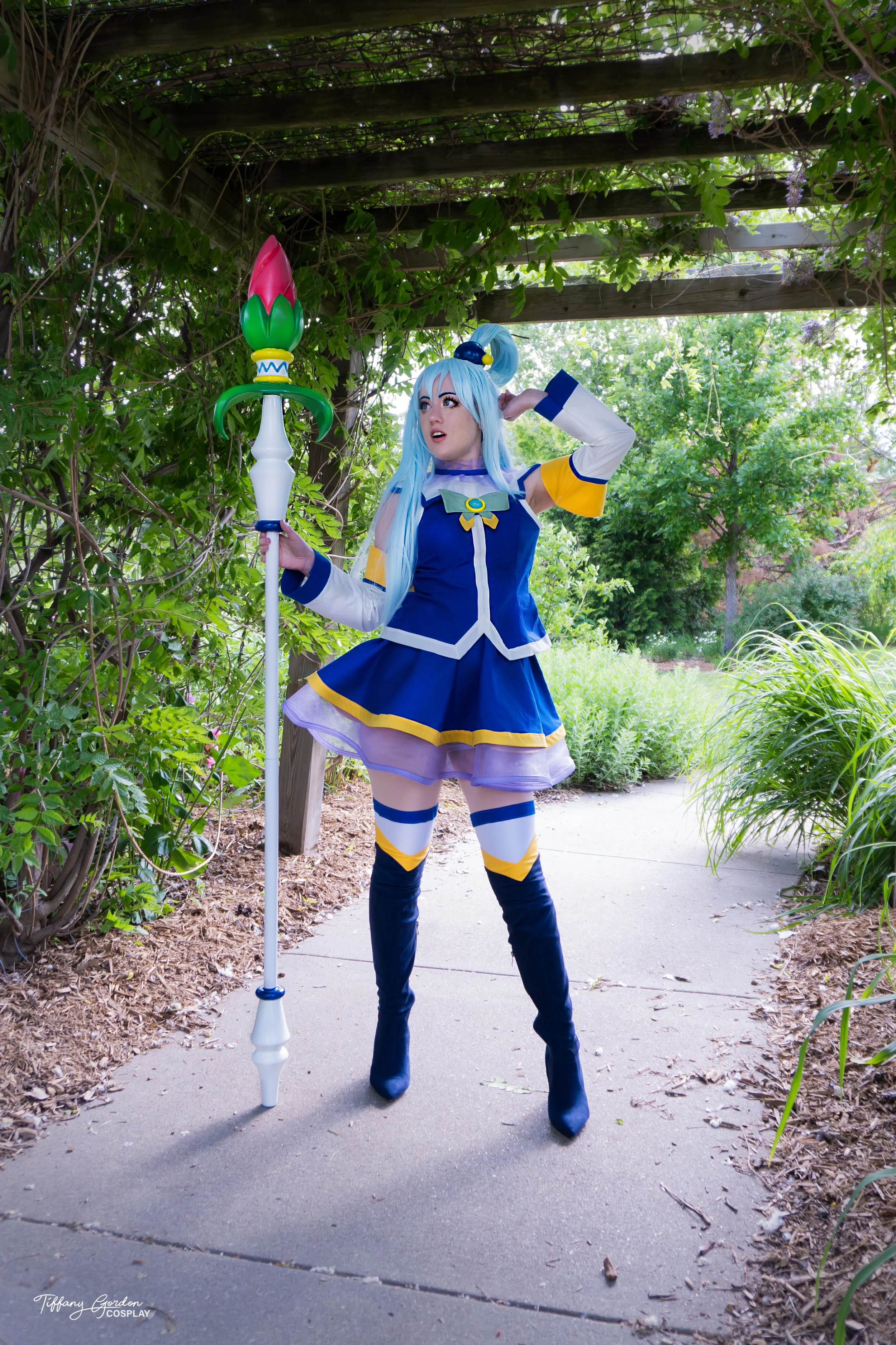 Aqua cosplay from KonoSuba by Tiffany Gordon Cosplay, featuring a blue wig, a blue and white dress with yellow bows, and a purple translucent divine ribbon holding a flower staff.