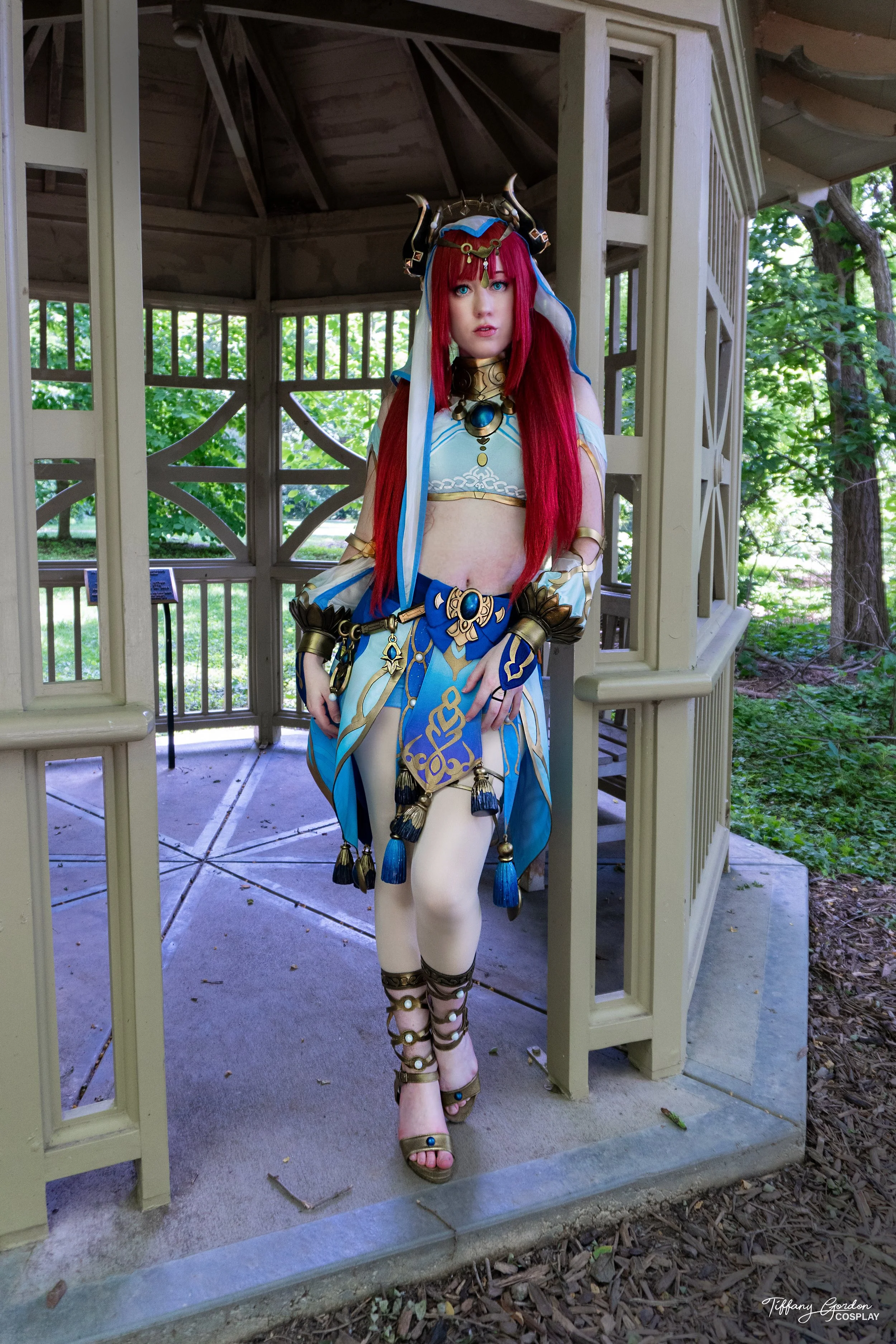 Tiffany Gordon Cosplay as Nilou from Genshin Impact. The cosplayer is standing in a wooden gazebo, wearing a highly detailed turquoise and white dancer’s outfit with gold trim, a red wig with black horns, and elaborate blue tassels.