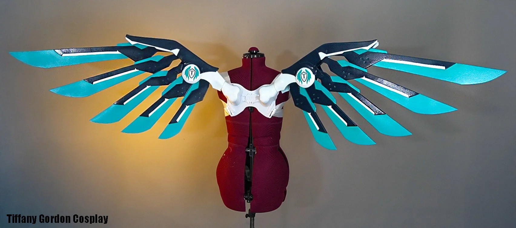 A technical display of a custom-built, detachable Valkyrie wing set for a Mercy cosplay by Tiffany Gordon Cosplay. The wings feature a blue and white mechanical frame with vibrant teal energy plumes and a central white harness mounted on a dress form