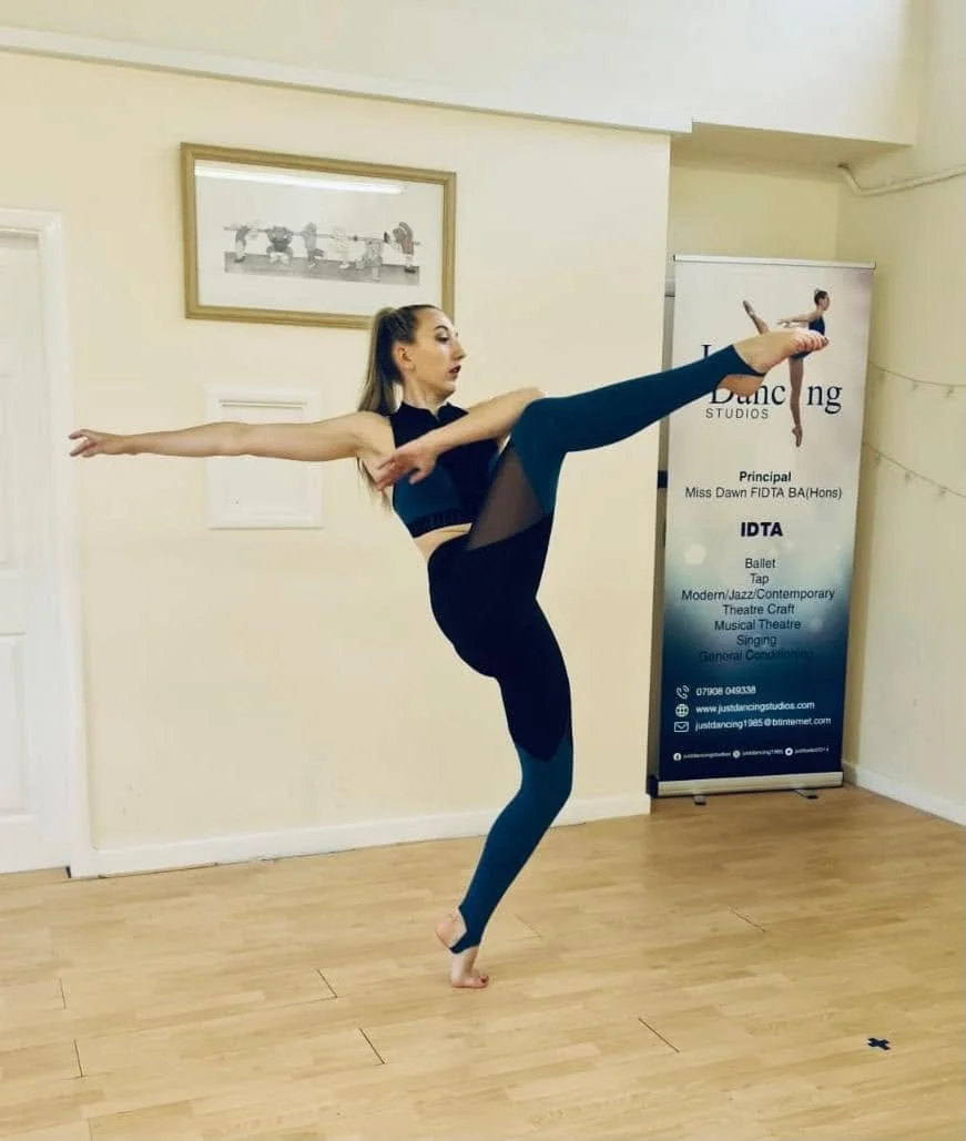 A young female dancer in black and teal exercise attire performs a high kick balancing on one foot in a dance studio.