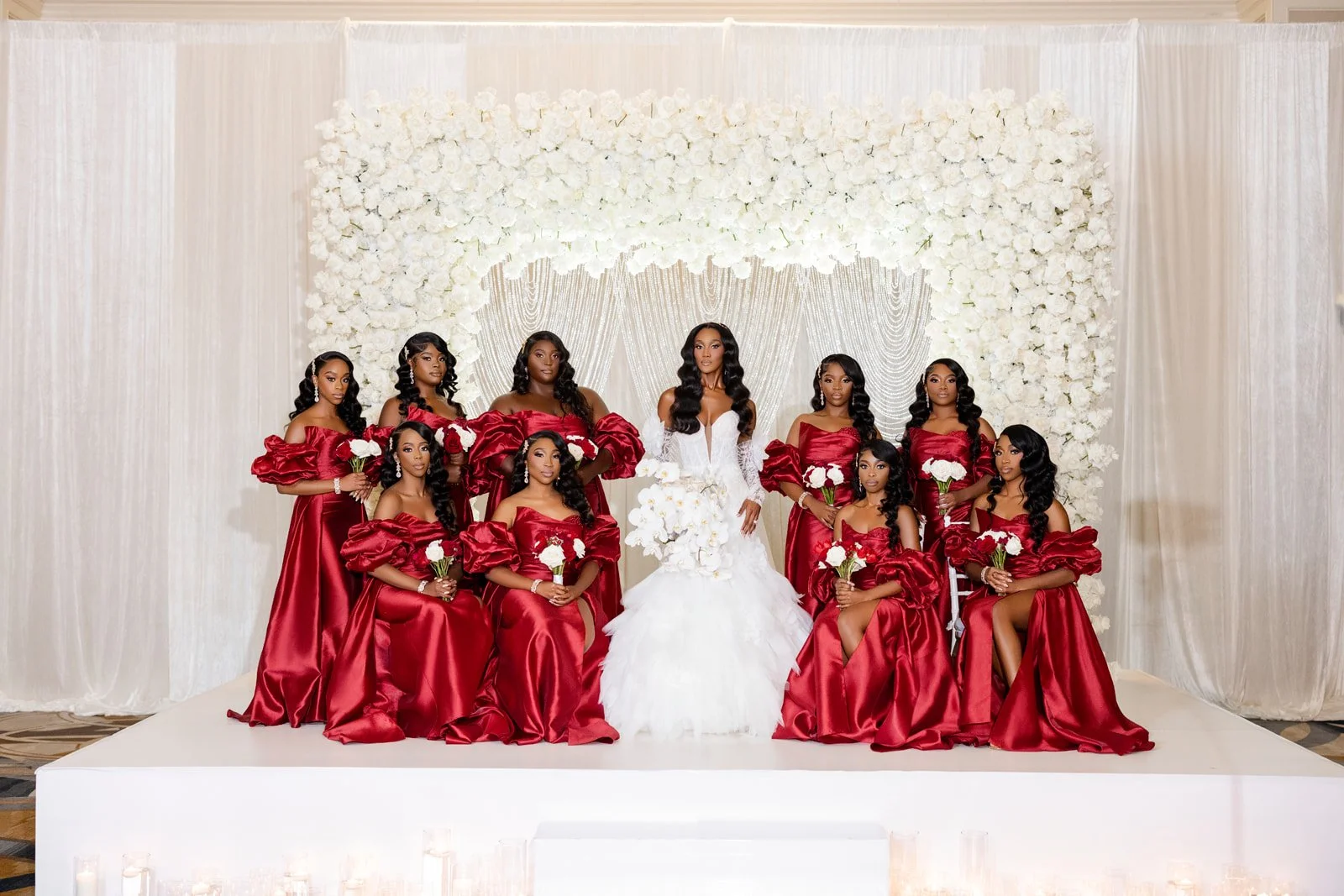 Bride with bridesmaids in red gowns before a white floral wall, photographed by Topflash Photography