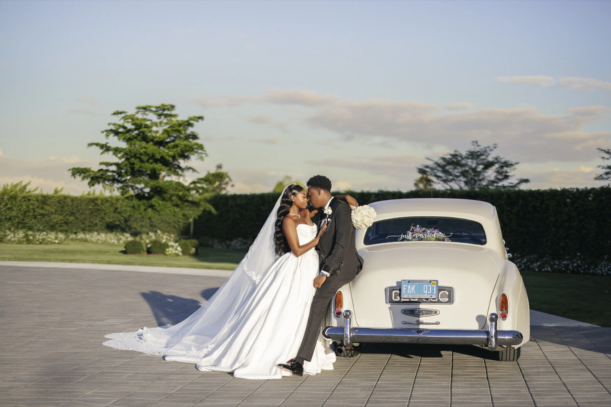 Jessica and Frantzdy kissing beside a classic cream vintage car in the late-afternoon light on their White Orchid wedding day