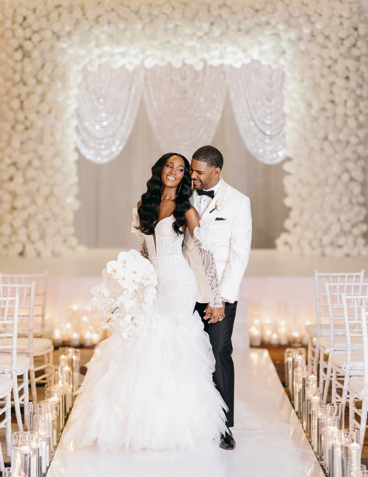 Bride and groom standing on a candlelit aisle before a white floral wall, photographed by Topflash Photography