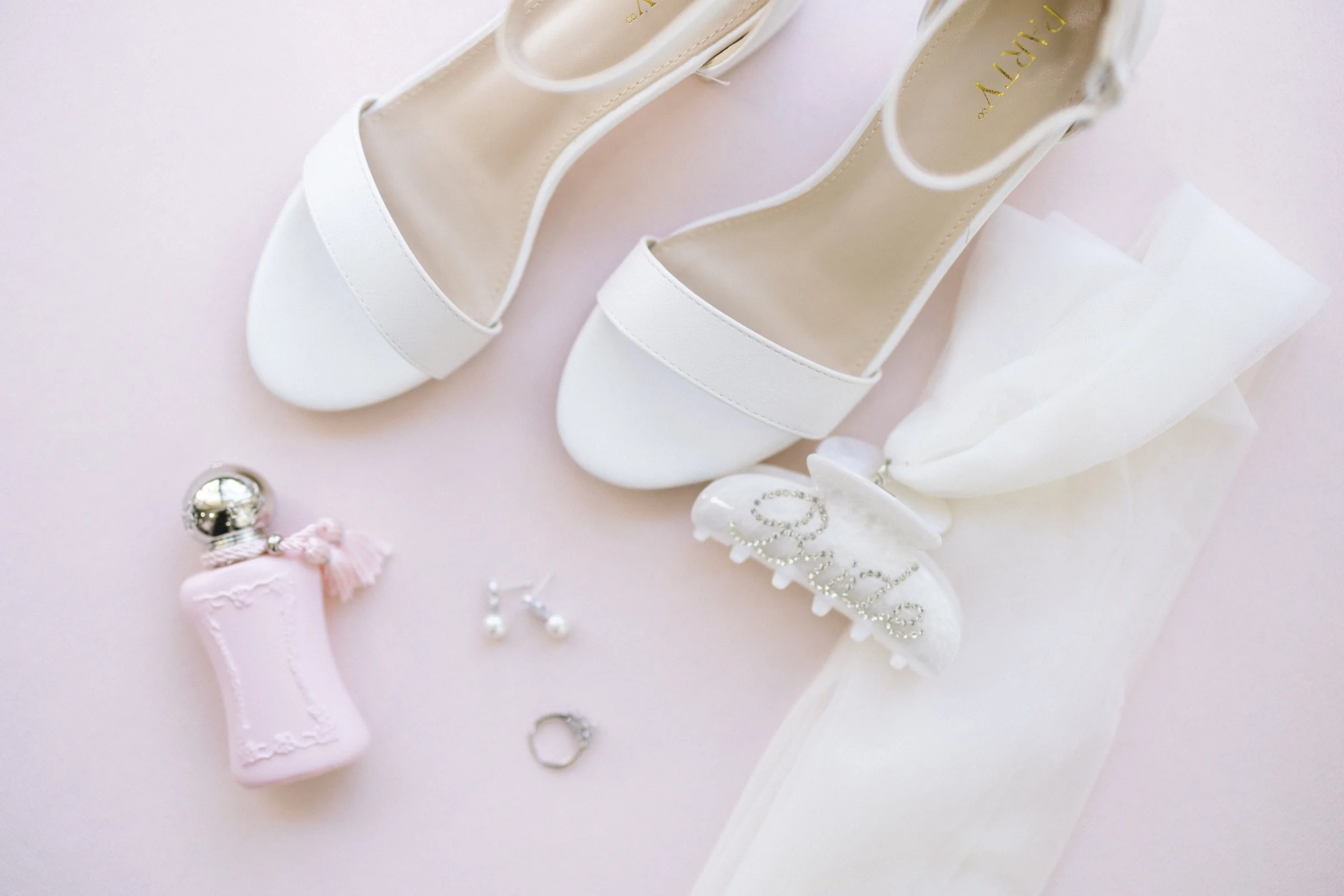 Bride's white strappy heels, wedding rings, pearl hair clip and pink perfume bottle styled on a blush backdrop at Lavan Venue in Hollywood, FL