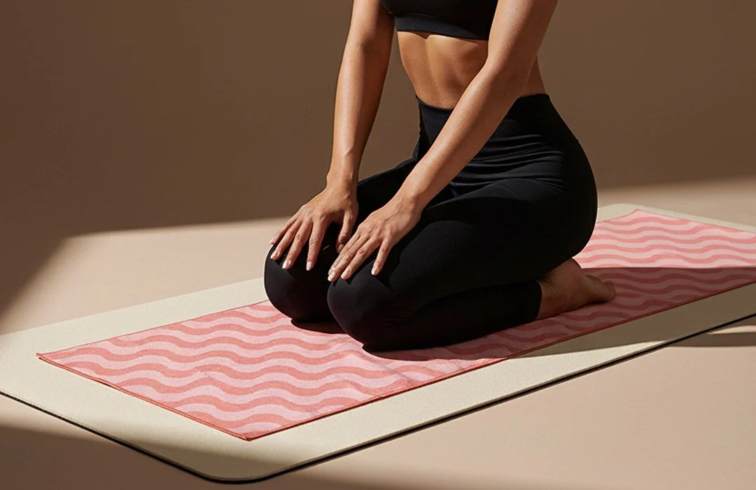 Woman kneeling on bikram, hot yoga towel on a yoga mat. She is in black yoga pants in a neutral coloured studio.
