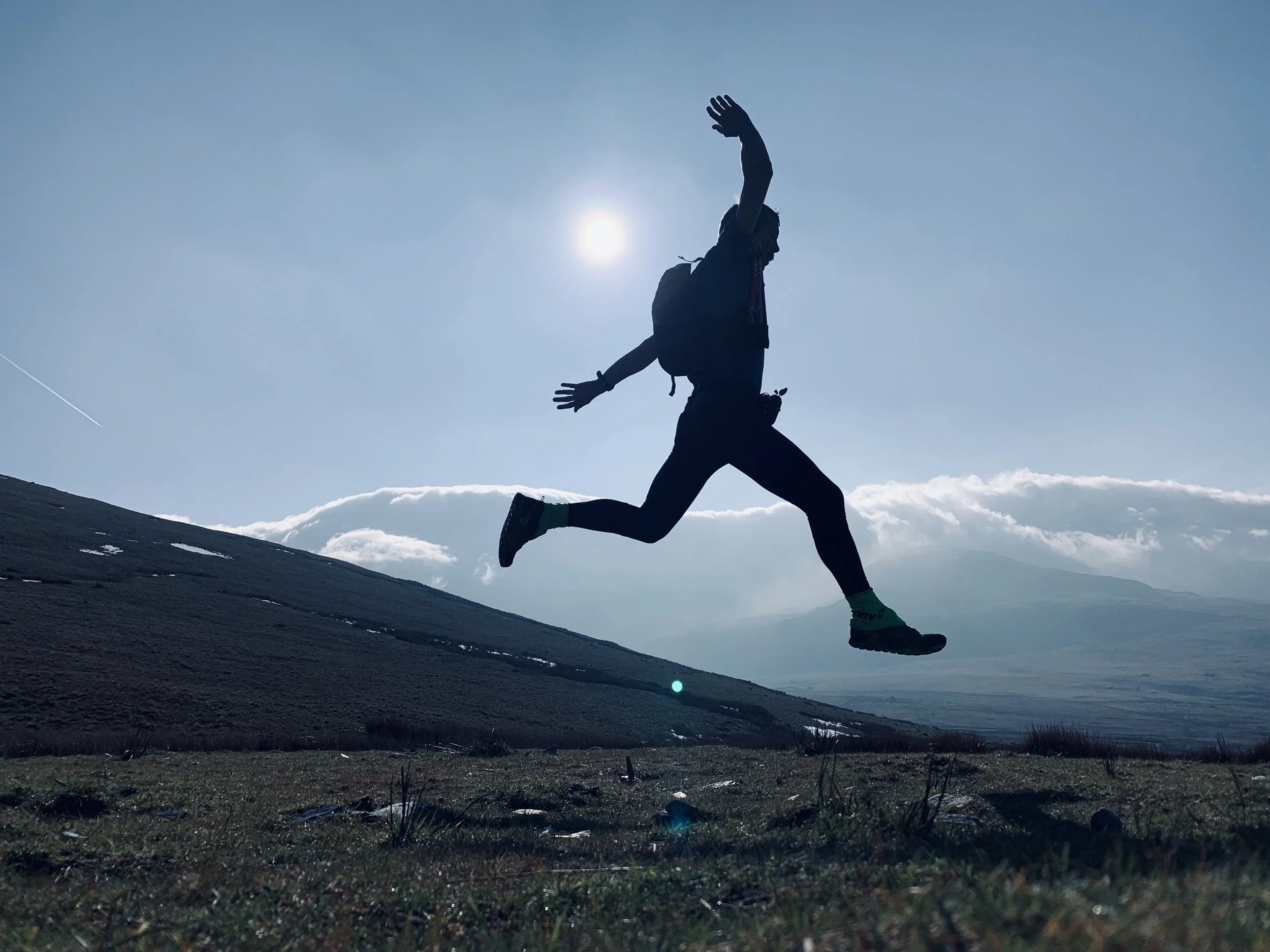 Kristian Morgan, running down a hillside with mountains and clouds in the background, with the sun behind them.