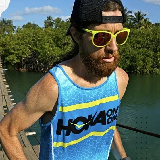 Man with long hair and beard wearing neon green sunglasses, a black cap backwards, and a blue athletic tank top with yellow and black text, standing on a wooden dock near water with greenery and trees in the background.