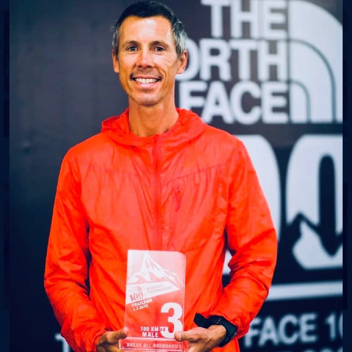 A man smiling and holding a red marathon bib in front of a black and white logo of The North Face. He is wearing a red jacket and a watch.
