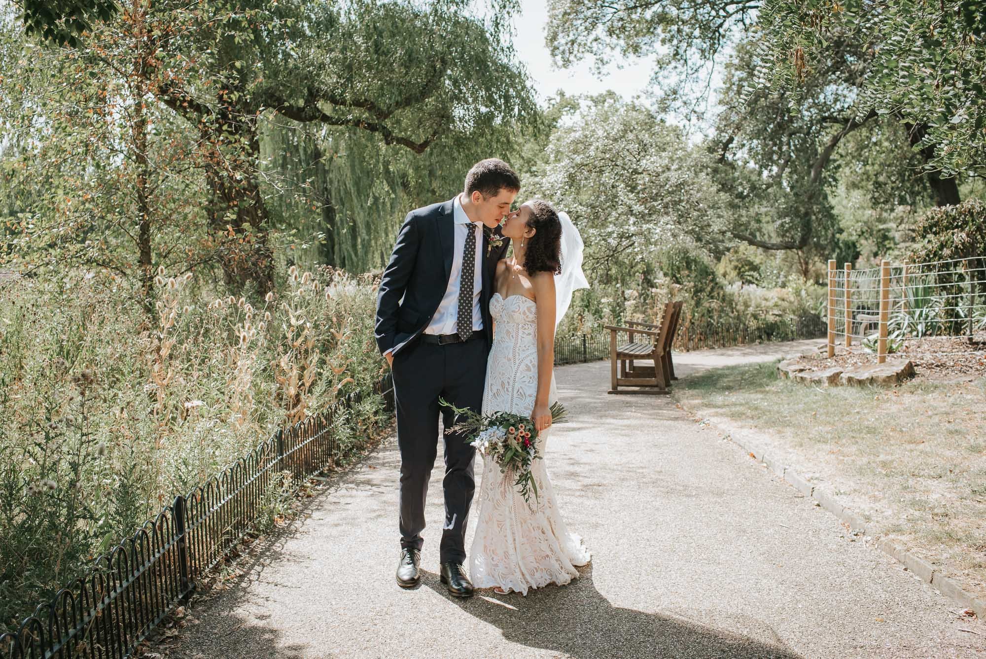  Bride and Groom walking hand in hand and kissing along a pathway in Regents Park in London. 