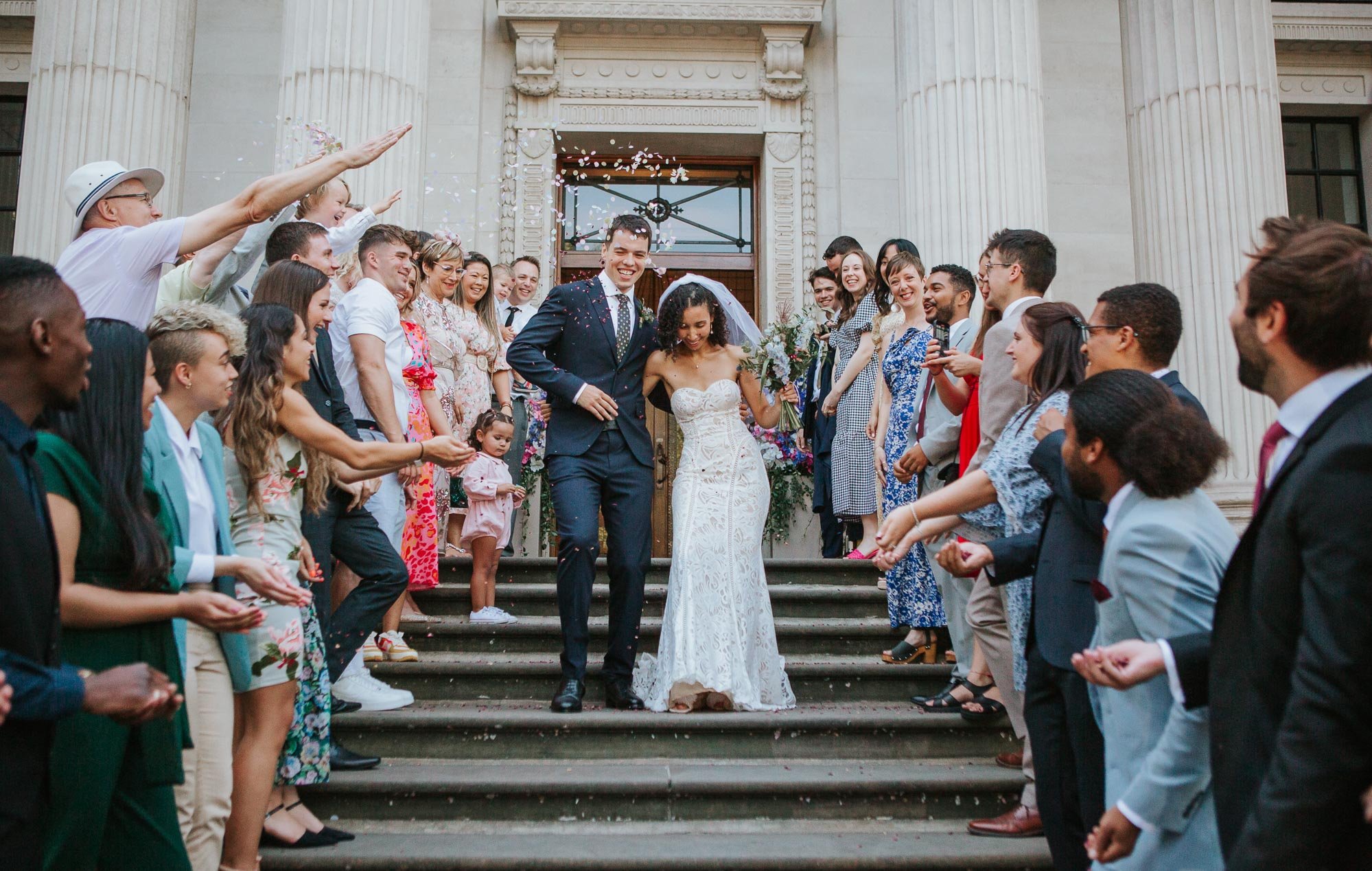  Groom and bride walking down the staircase, with confetti being thrown at them, outside Marylebone Town Hall in Marylebone, London.   