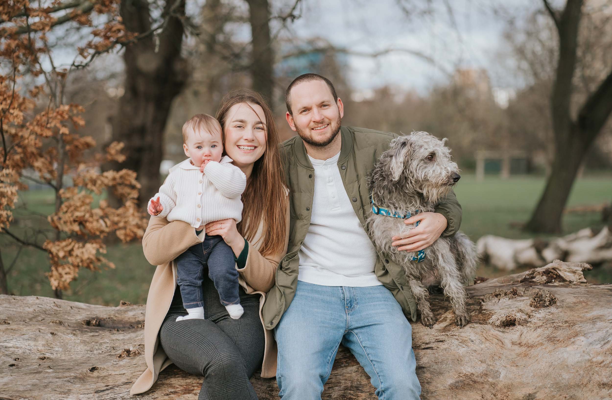  Family of mum dad , son and dog sitting on a log in Brockwell Park posing for photographer christine constantine photography. 