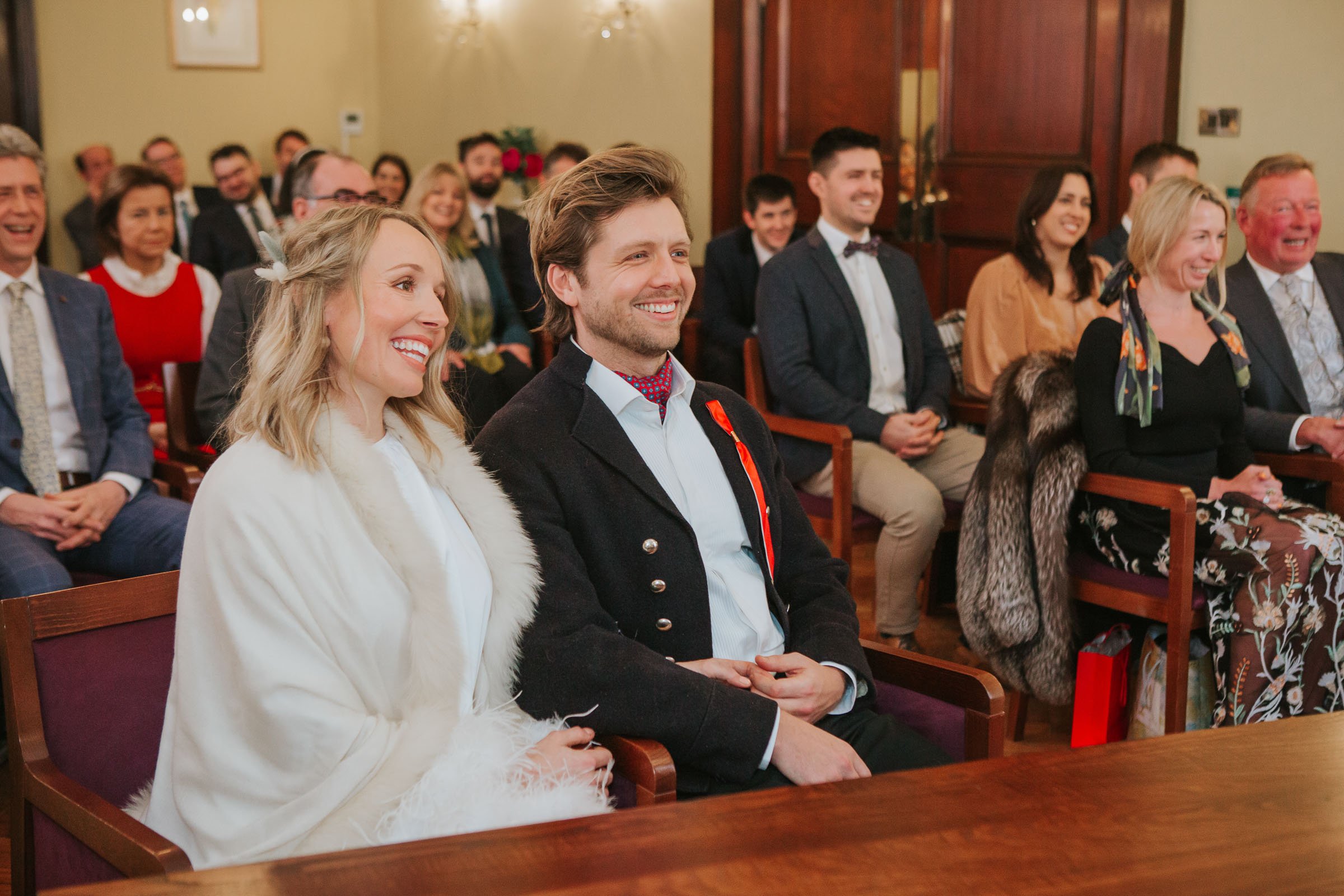  Lizzie and Max sitting next to each other in front of their friends and family in The Byron Room at Chelsea Old Town Hall.  