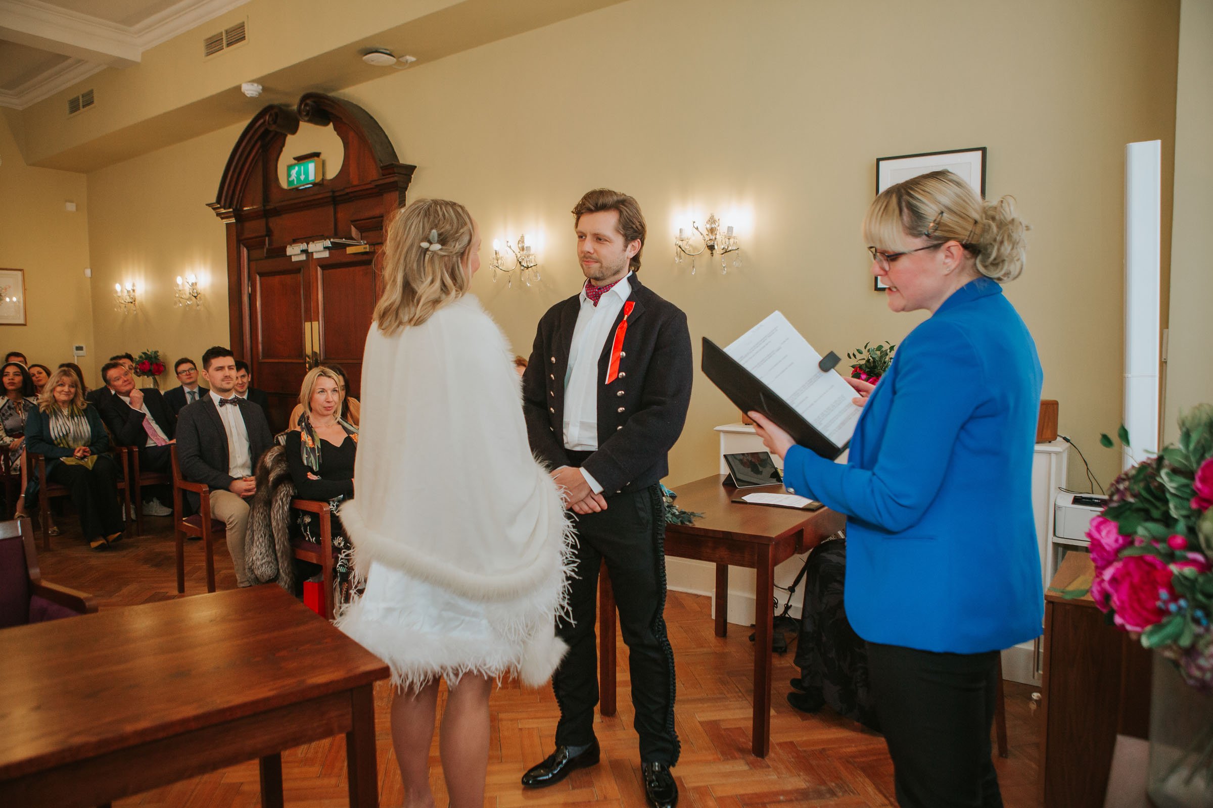  Lizzie and Max holding each others hands and looking into each others eyes in front of their friends and family in The Byron Room at Chelsea Old Town Hall. 