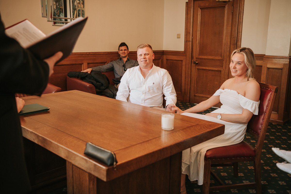  Steven and Sarah sitting down hand in hand looking at the registrar while exchanging vows at their wedding at The Elizabeth Room at Wandsworth Town Hall Registry Office. 