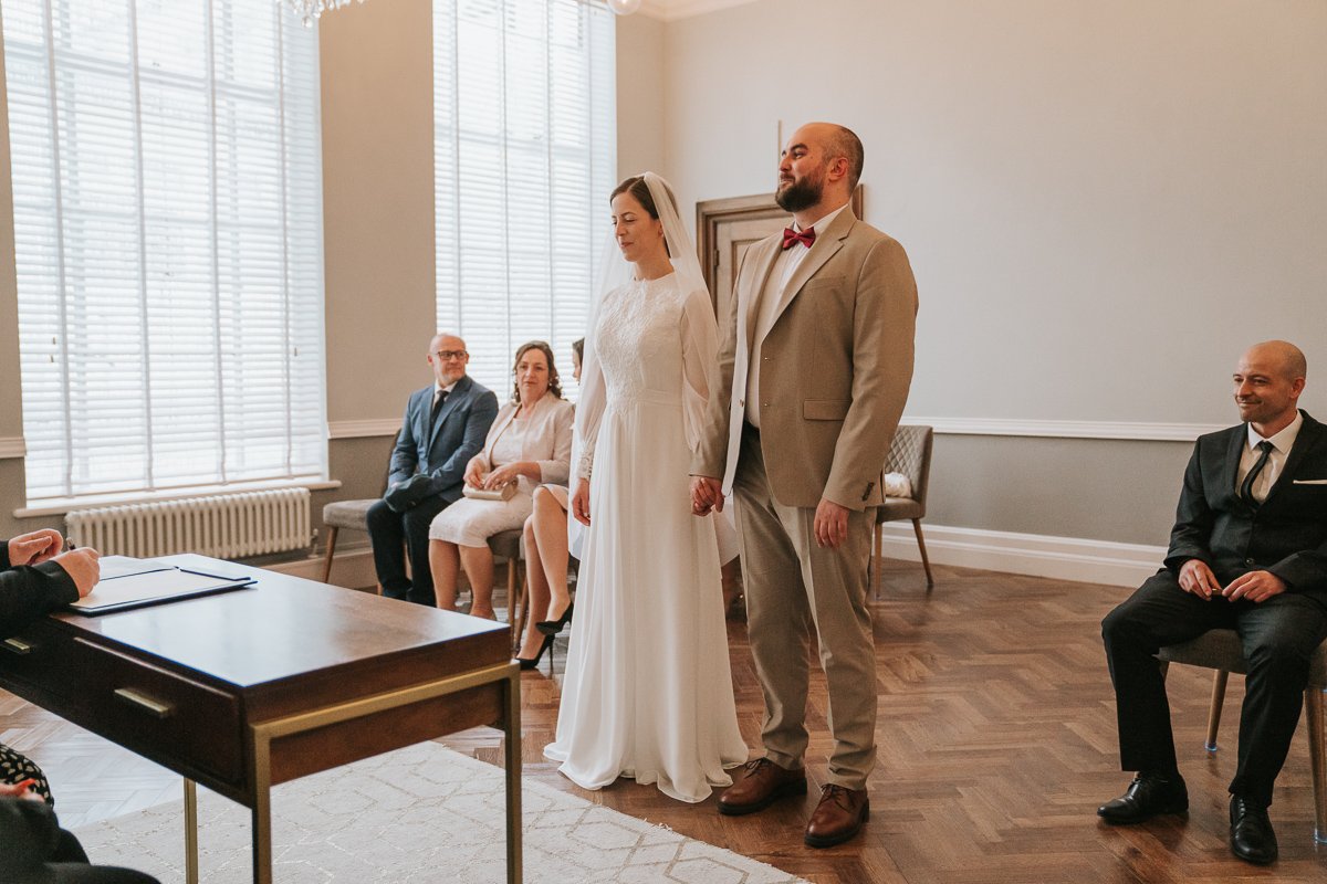  Edina and Zsolt, the bride and groom, stand next to each other in the wedding ceremony room at Islington Town Hall. 