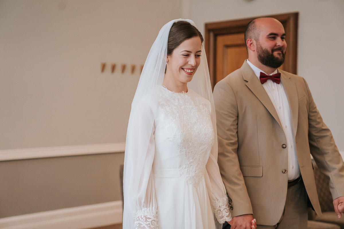  Edina and Zsolt, the bride and groom, stand next to each other smilng as they get married in the wedding ceremony room at Islington Town Hall. 