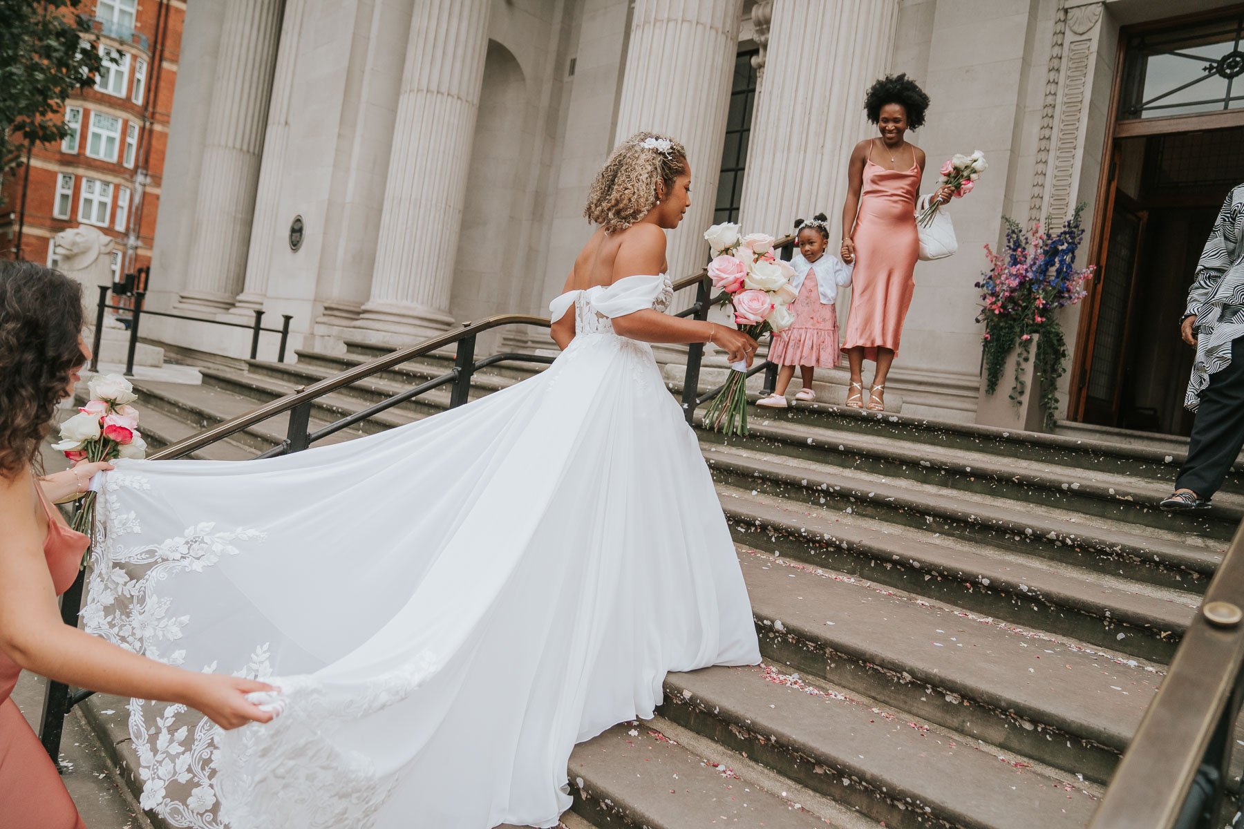  Lucia walks up the steps of Marylebone Old Town Hall with her dress flowing and held by her bridesmaids 