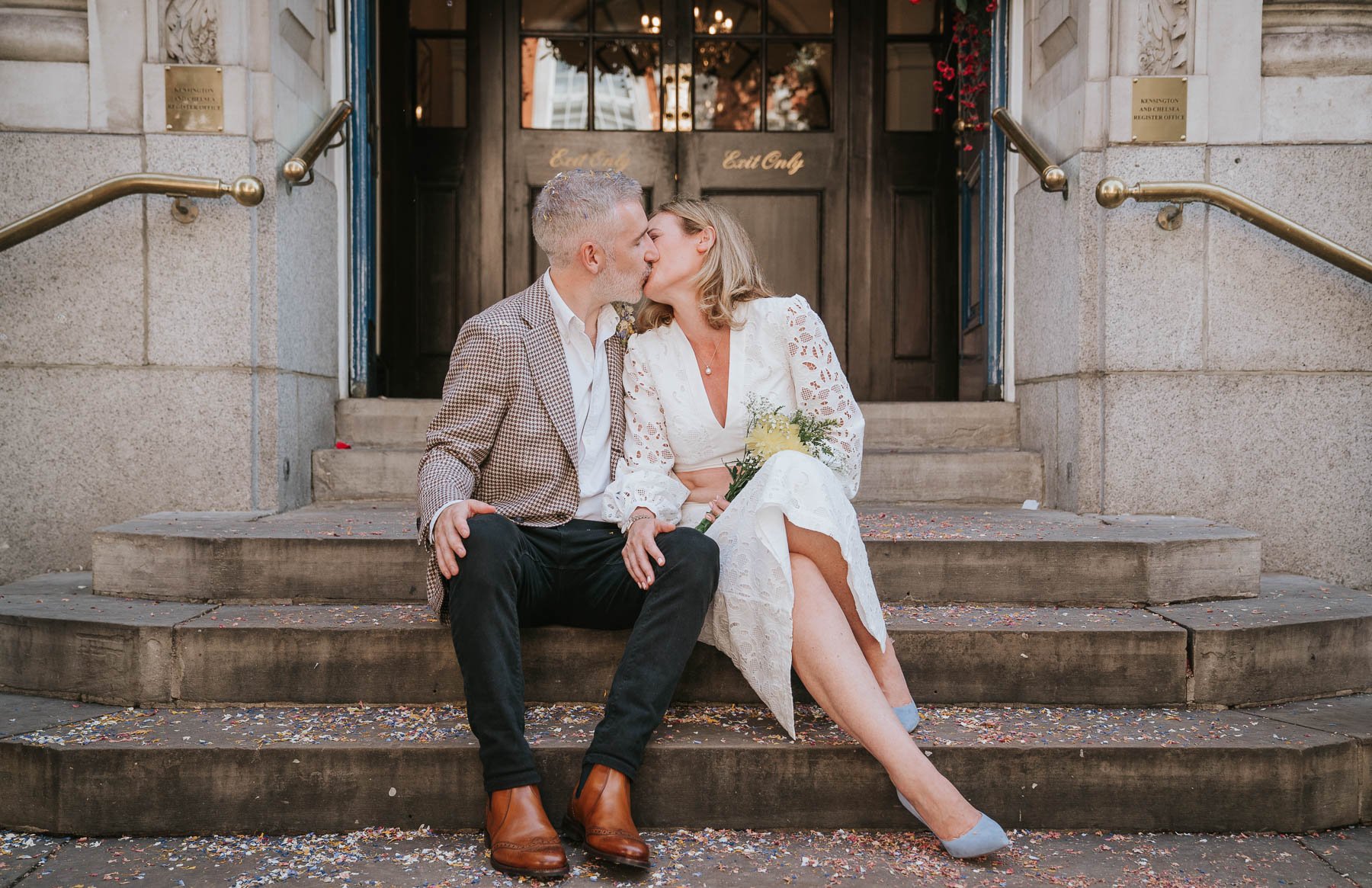  Bride and groom sit and kiss on steps outside Chelsea Old Town Hall. 