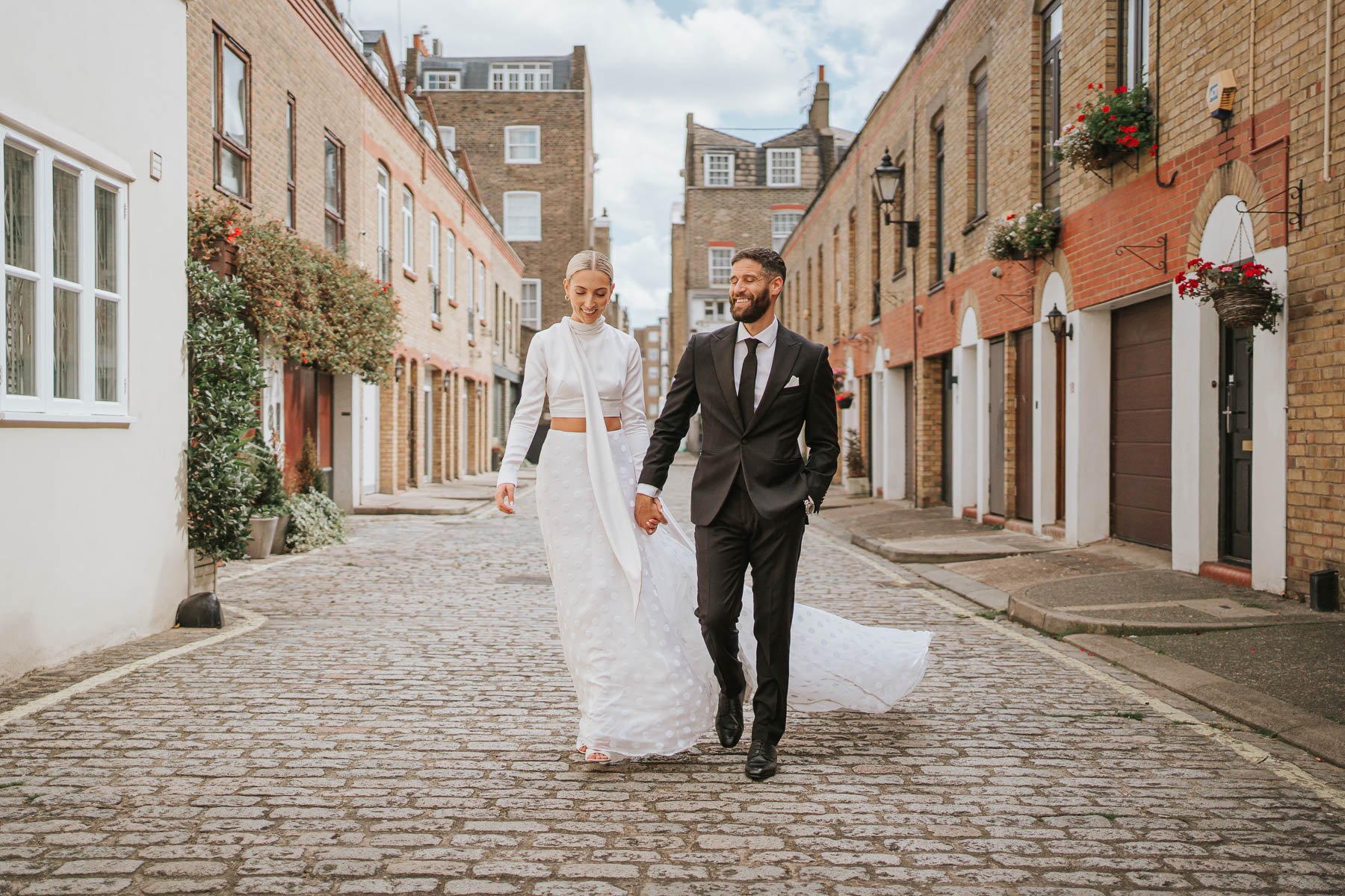  Bride and groom walk towards photographer arm in arm down cobbled street behind Marylebone Town Hall.  