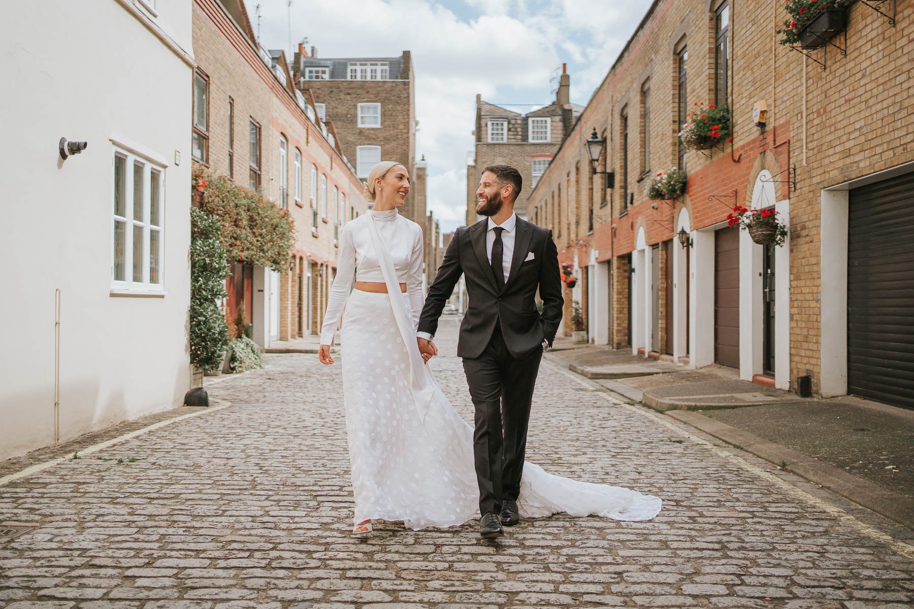  Bride and groom walk towards photographer hand in hand down cobbled street behind Marylebone Town Hall.  