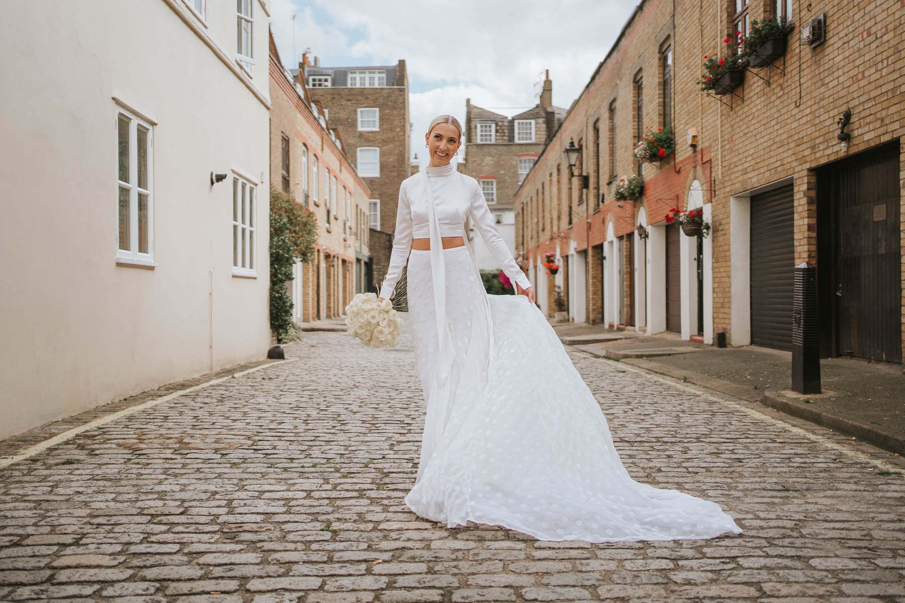  Bride poses with flowing wedding dress in middle of cobbled street behind Marylebone Town Hall. 