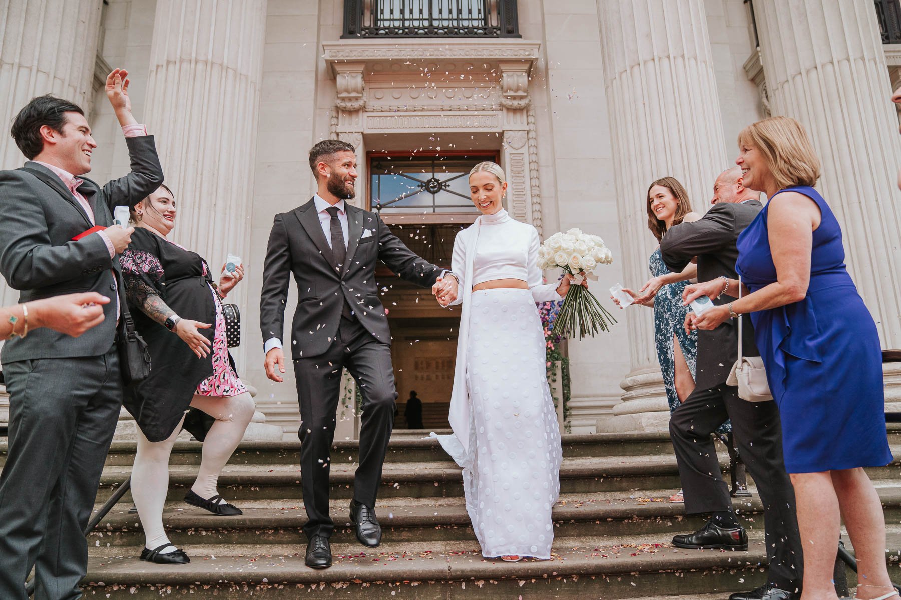  Bride and groom walk down steps outside Marylebone Town Hall while being showered in confetti. 