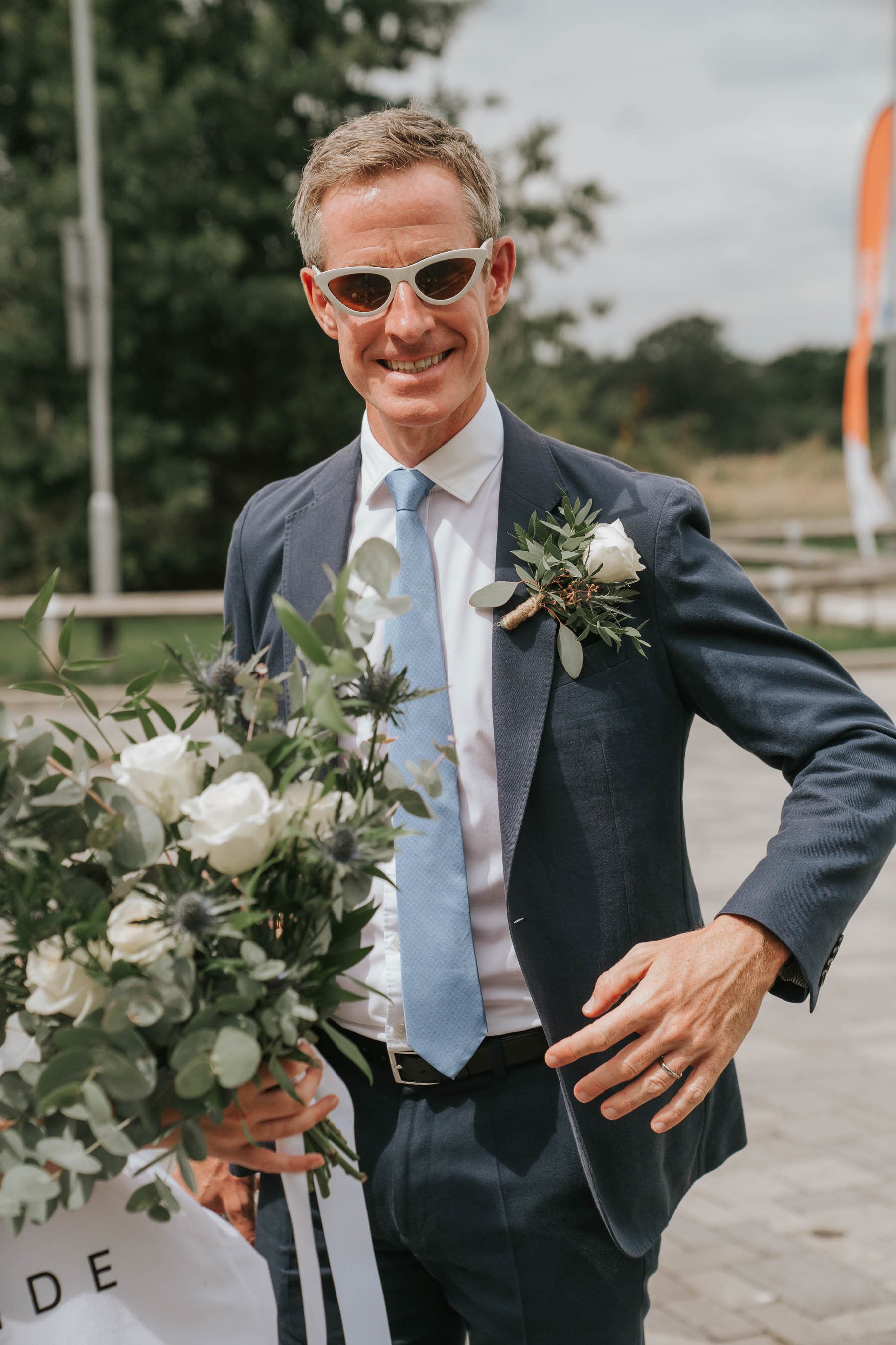  Groom wearing bride’s sunglasses and holding flowers in Morden Park House car park. 