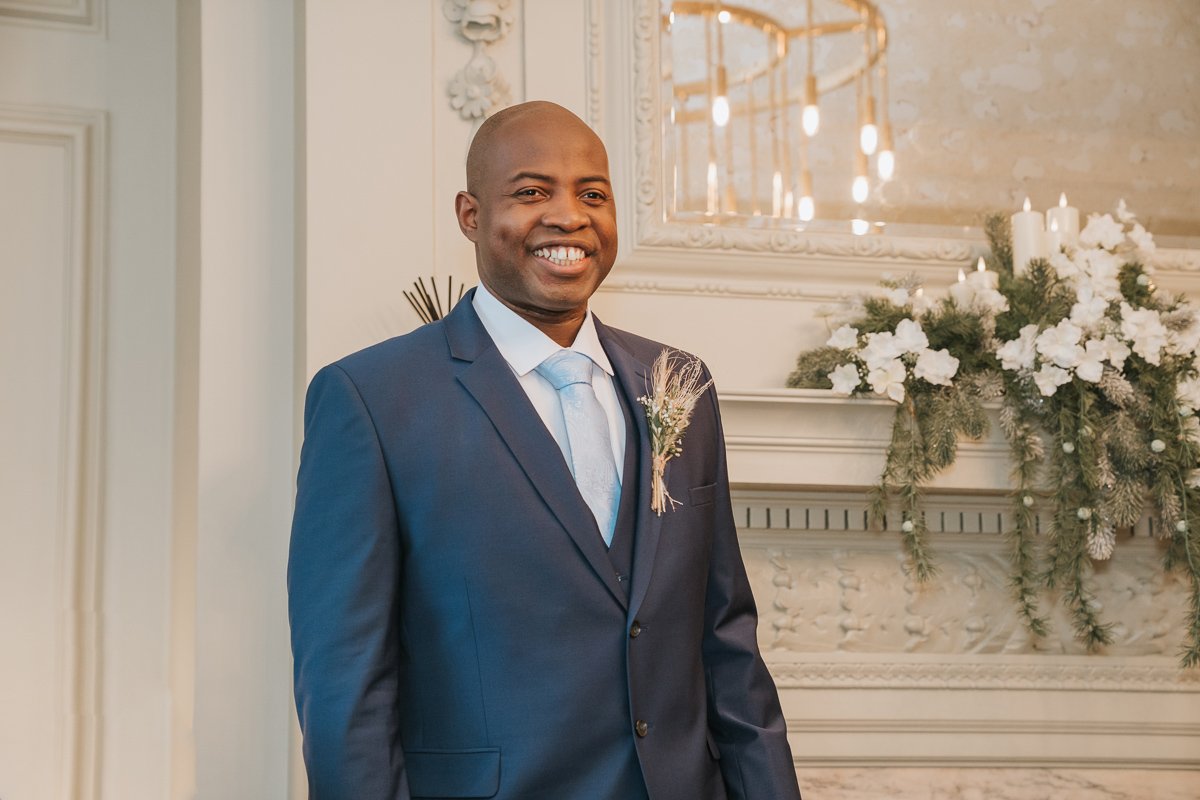  A groom stands smiling in front of the fireplace in the pimlico room at marylebone town hall. 