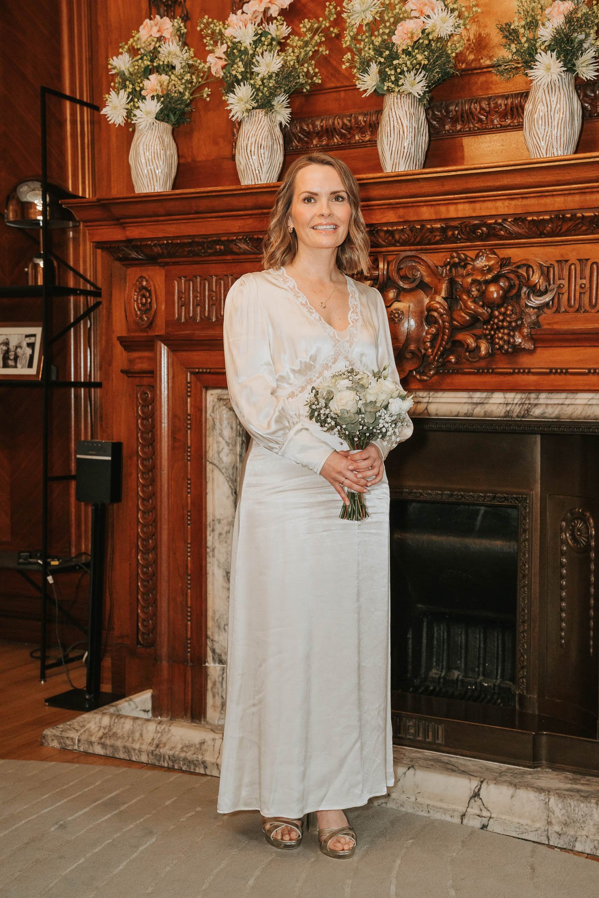  Bride standing in front of fireplace holding wedding flowers, in the Paddington Room at Old Marylebone Town Hall. 