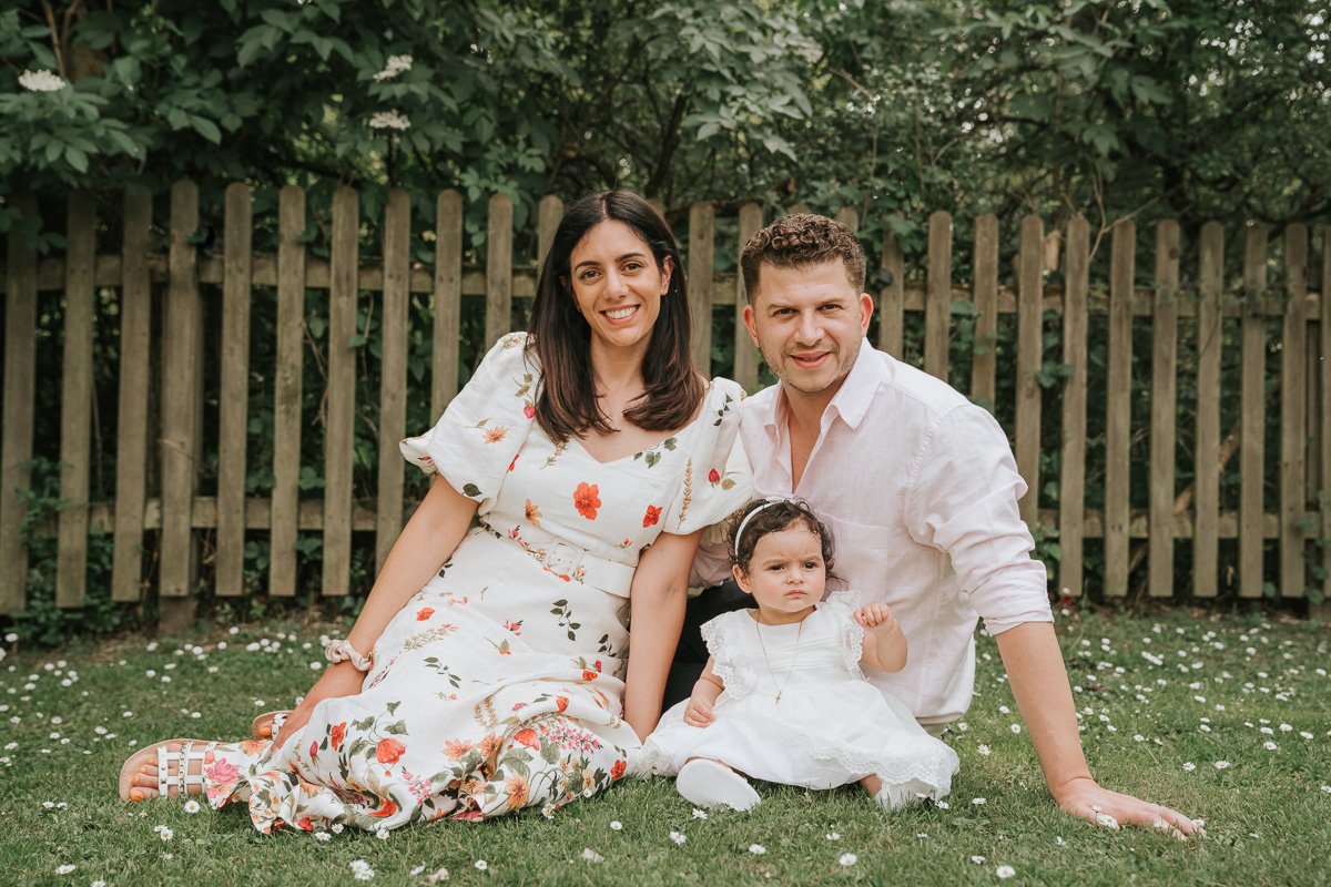  Mum, Dad and newly-Christened baby girl sit together on grass and pose for a photograph. 