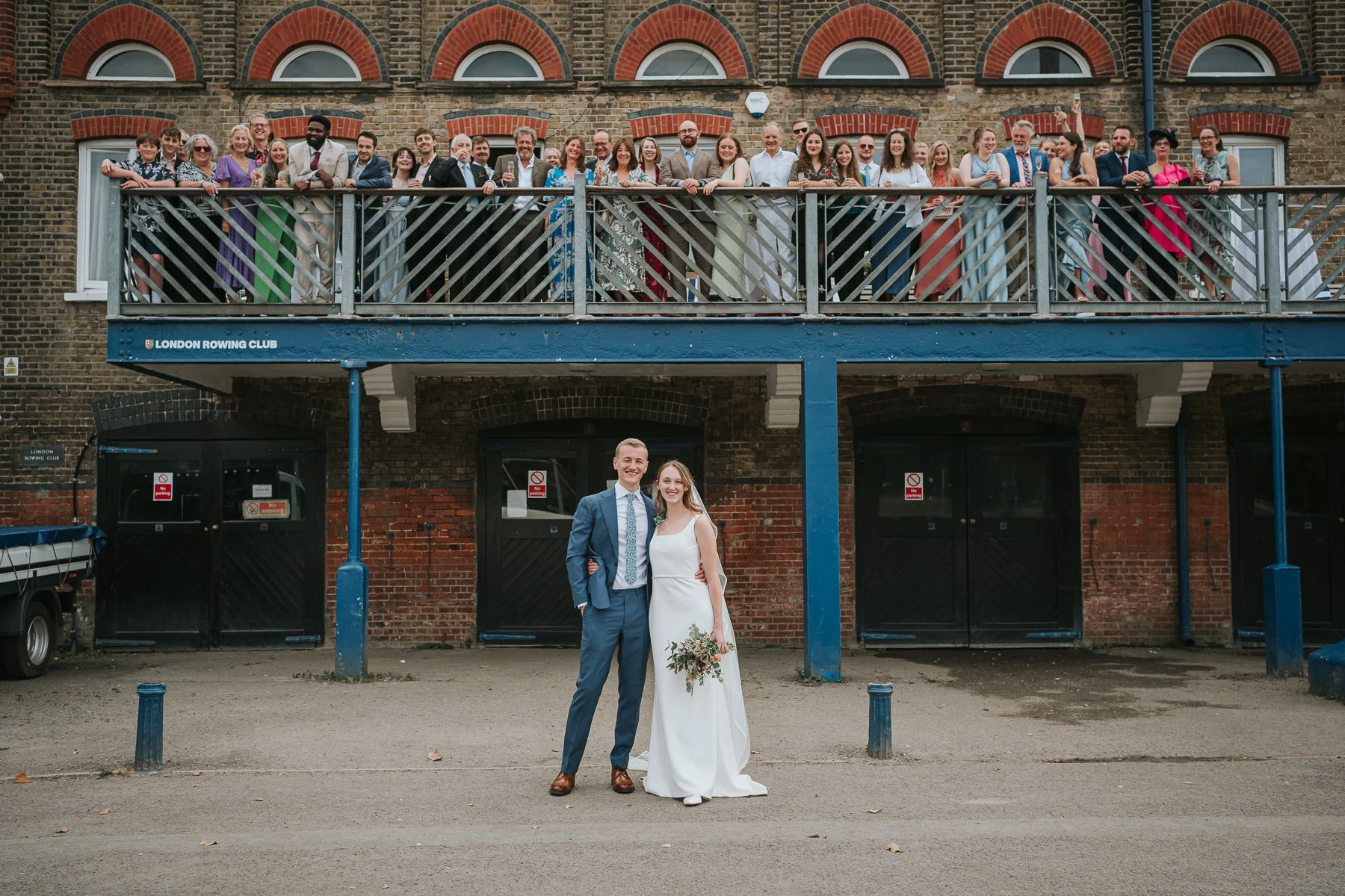  Couple pose for photograph after their wedding in London Rowing Club. Friends and family look down from balcony.  