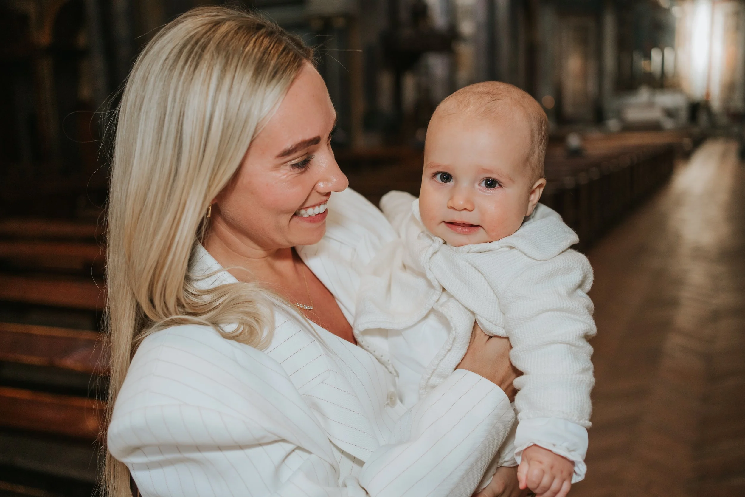  Mother holding her baby in a white christening outfit inside Brompton Oratory, London, with warm light highlighting the church’s ornate interior 